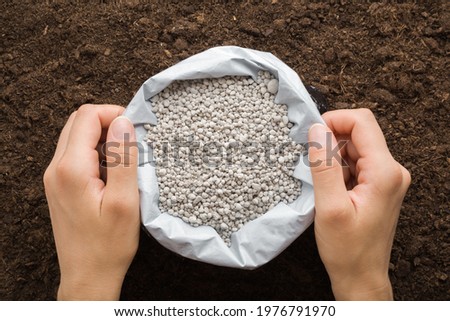 Young woman hands holding opened plastic bag with gray complex fertiliser granules on dark soil background. Closeup. Point of view shot. Product for root feeding of vegetable, flowers and plants.  Royalty-Free Stock Photo #1976791970
