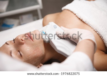 Woman client lying in beauty salon room, receives ultrasound smas non-surgical face lift by professional beautician doctor, hand holding ultrasonic hardware cosmetology device for facial treatment Royalty-Free Stock Photo #1976822075