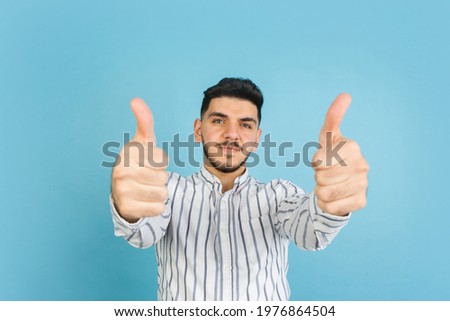 Thumbs up. Latino young man's portrait on blue studio background. Handsome male model in casual style. Concept of human emotions, facial expression, youth, sales. Copy space for ad.