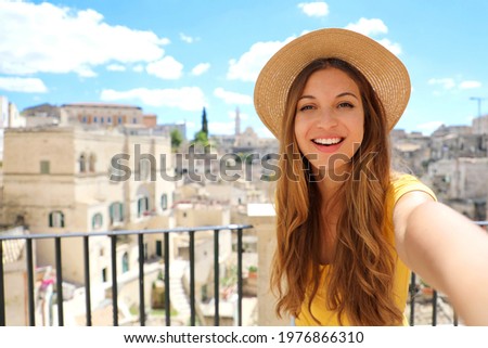 Happy beautiful female tourist is taking a selfie smiling at the camera with the city of Matera on the background in Italy