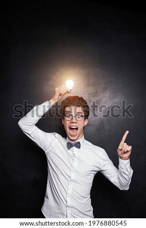 Funny young male genius in nerdy glasses and bow tie with shiny light bulb over head pointing up, while representing business idea concept against black background