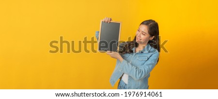 panoramic banner. happy asian pretty woman in casual clothing smile, hold empty blank blackboard with copy space area for text on yellow background, online marketing, advertising, announcement concept