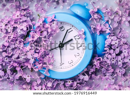 clock alarm clock is buried in lilac flowers, daylight savings time, spring time Royalty-Free Stock Photo #1976916449
