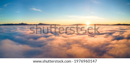 Aerial view of yellow sunset over white puffy clouds with distant mountains on horizon. Royalty-Free Stock Photo #1976960147