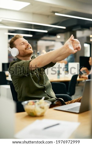 Happy businessman enjoying in music over headphones and stretching after working on laptop in the office.