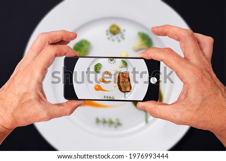 HANDS TAKING A CULINARY PHOTOGRAPH WITH A SMARTPHONE TO A GOURMET AND MINIMALIST SALMON DISH. CREATIVE HIGH CUISINE MICHELIN STAR. KETO DIET RECIPES. TOP VIEW.