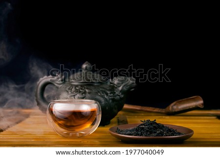 Chinese tea ceremony. Clay teapot with red tea Lapsang souchong on a black background, heap of dry tea leaves and glass thermo cup of hot tea with vapour.