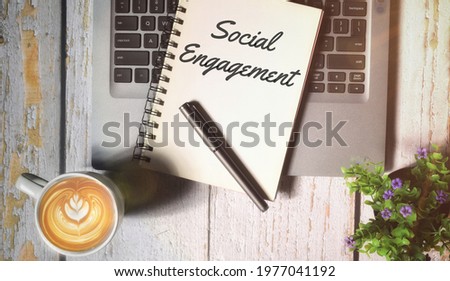 Top view of a cup of coffee, book, pen and laptop with Social Engagement wording over a wooden background with vintage and flare effect. Selective focus image