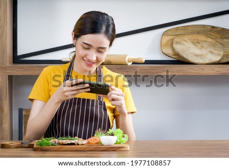 Young asian housewife dressed in an apron, using mobile phone to take pictures of delicious steaks. Morning atmosphere in a modern kitchen.