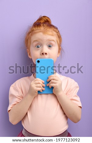 surprised redhead child girl using smartphone, isolated on purple background in studio. checking news, surf the net, look at camera with big blue eyes, closing face with blue mobile phone