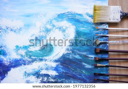 Painter paint acrylic on canvas with brush with blue ocean on frame. Artwork as hobby or occupation for artist. Big wave in sea w creates artwork by mix color and decors picture as hard copy art.