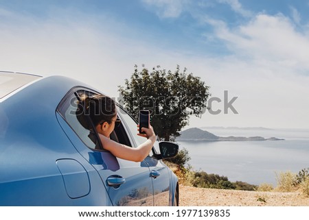 Young woman taking picture with smartphone from open window of a car stopped roadside with a beautiful view to the coast