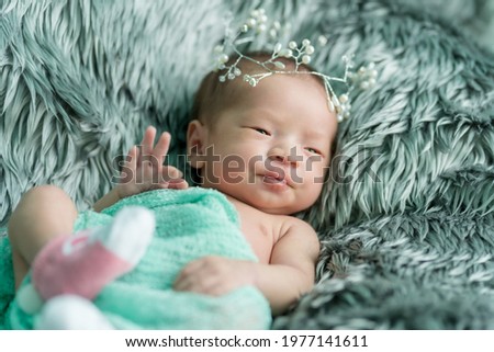 A newborn baby wrapped in a green cloth with crown was placed on a gray fluffy rug. She was lying facing to the right of the picture and raised her hand, gesturing okay.