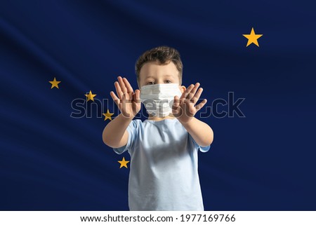 Little white boy in a protective mask on the background of the flag of Alaska. Makes a stop sign with his hands, stay at home Alaska.