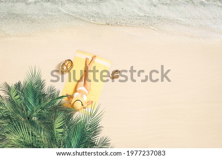 Alone woman in straw hat and bikini sunbathes on yellow beach towel under palm tree branches. Female relaxation on the sand beach at summer vacation. Top view
