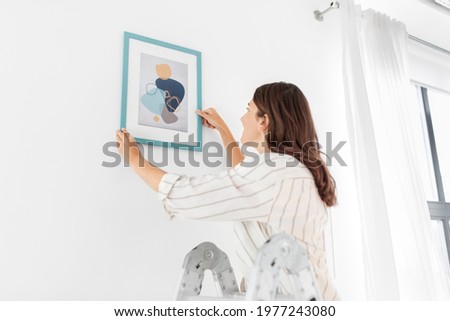 home improvement, decoration and people concept - happy smiling woman on ladder decorating home with art