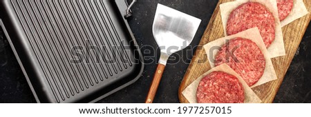 Griddle Grill Pap and Ground Beef Meat Patties for Grilling on Black Background, Overhead View. Raw Steak Burgers Cutlets On Grill Pan.  Burgers for Frying, Grill Tools and Cutting Board, Top View.
