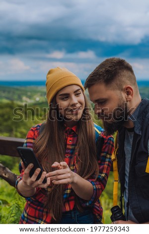 A young couple of hikers using smartphone for orientation while spending time in nature and standing on the lookout with camping backpacks