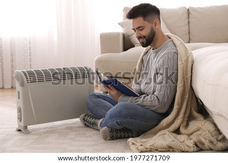 Young man warming up near electric heater at home Royalty-Free Stock Photo #1977271709