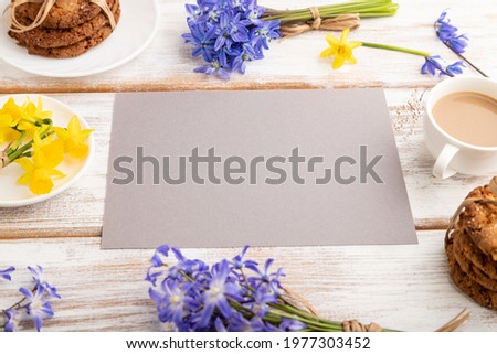 Gray paper sheet with oatmeal cookies, spring snowdrop flowers bluebells, narcissus and cup of coffee on white wooden background. side view, copy space, still life. Breakfast, morning, spring concept.