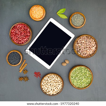 Flat lay of various sources of vegetable protein: beans, lentils, peas, chickpeas, mung bean in bowls. Tablet with black screen. Black background. Top view, copy space. Online concept. 