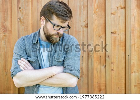 Pensive, attractive bearded man with glasses crossed his arms and looks down, against a background of a wooden wall. Copy space.