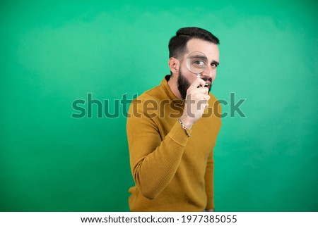 Handsome man wearing casual clothes over isolated green background Handsome man wearing casual clothes over isolated green background surprised looking through a magnifying glass