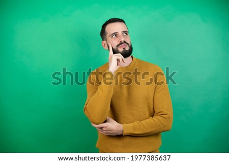 Handsome man wearing casual clothes over isolated green background with serious face thinking about question with hand on chin, thoughtful about confusing idea