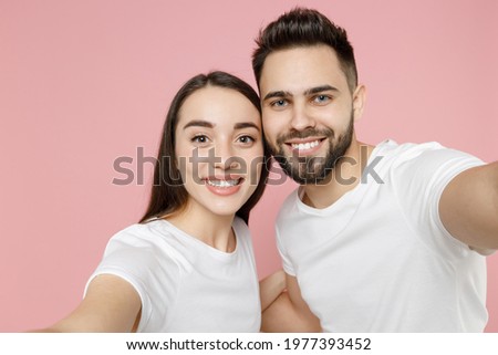 Close up young cheerful couple two friends man brunette woman in white basic blank print design t-shirts doing selfie shot on mobile cell phone isolated on pastel pink color background studio portrait