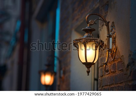 Lantern with yellow warm light on facade of building. Twilight on city street, building illumination. Retro lantern lighting, warm light glow. Street lights, illumination and vintage lantern Royalty-Free Stock Photo #1977429065