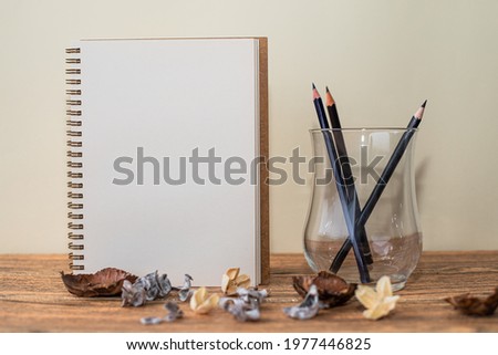 Open spiral notebook with white sheet, pencils in the glass next to it and decorations on the table