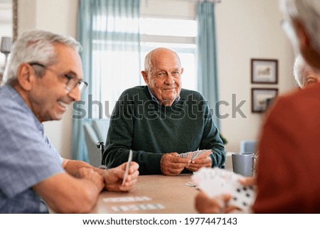 Retired senior man in wheelchair playing cards with friends at nursing home. Smiling group of old people playing cards at care centre. Happy seniors playing at lunch table and having fun together. Royalty-Free Stock Photo #1977447143