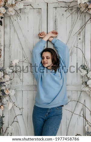 A girl with closed eyes leans against a white door decorated with flowers. Her arms are raised above her head and crossed. The girl is dressed in a blue sweater and blue jeans. She smiled fondly.