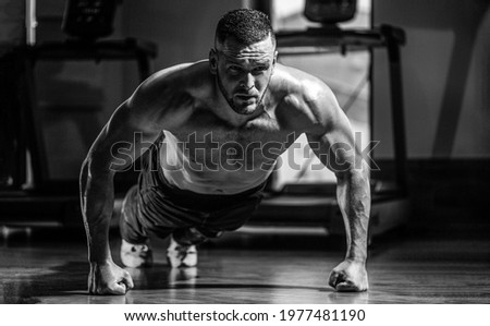 Muscular man doing push-ups on one hand against gym background. Man doing push-ups. Muscular and strong guy exercising. Slim man doing some push ups a the gym. Black and white. Royalty-Free Stock Photo #1977481190