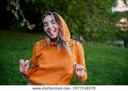 A young European happy woman with closed eyes wearing orange hoodie looking to the right. A fun picture with a model showing her tongue. Bright colourful photo. Copy space for text.