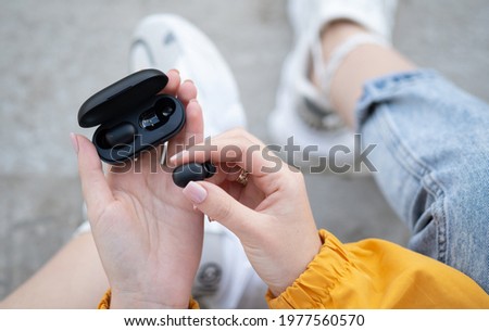 close-up of a woman taking out a black wireless earbud from his charging box. Female hands touching a portable gadget headphones Royalty-Free Stock Photo #1977560570