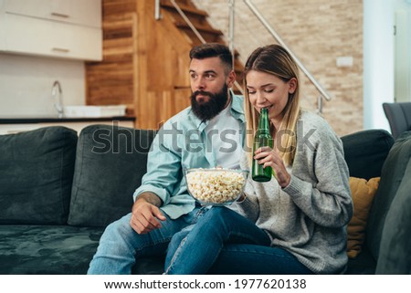 Young beautiful couple drinking beer and eating popcorn while watching television together at home