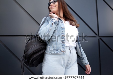 Fashion oversize woman, dressed white t-shirt , bagpack, sunglasses and jeans posing against the wall
