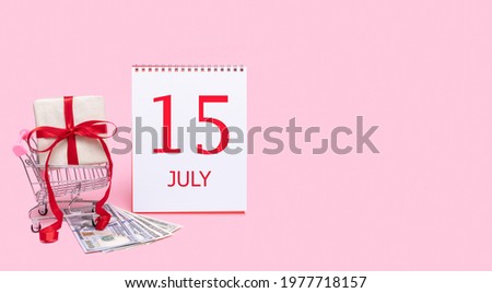 A gift box in a shopping trolley, dollars and a calendar with the date of 15 july on a pink background.