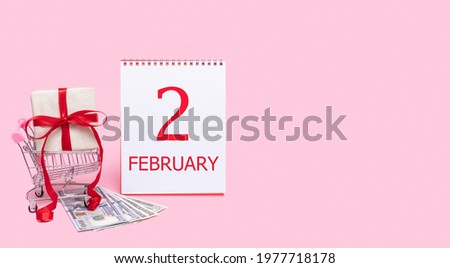 A gift box in a shopping trolley, dollars and a calendar with the date of 2 february on a pink background.