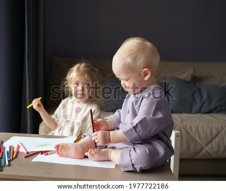 Cute baby boy with golden hair sitting on table, playing with colorful pencils while his older sister creative little girl drawing something on piece of paper. Children and art activities at home