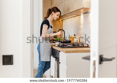 Young woman in apron cooking healthy food at modern home kitchen. Preparing meal with frying pan on gas stove. Concept of domestic lifestyle, happy housewife leisure and culinary hobby. Royalty-Free Stock Photo #1977760286