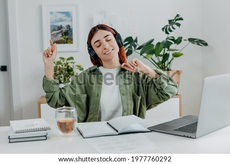 Woman listening music with wireless headphones. Young businesswoman relaxing at comfortable workplace. Concept of resting during online business at home office,telework or studing. Happy girl singing. Royalty-Free Stock Photo #1977760292