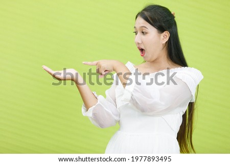 Image is portrait. Asian smiling plump woman wearing white dress. Lady asia standing use her index finger on  hand and shocked expression in green background