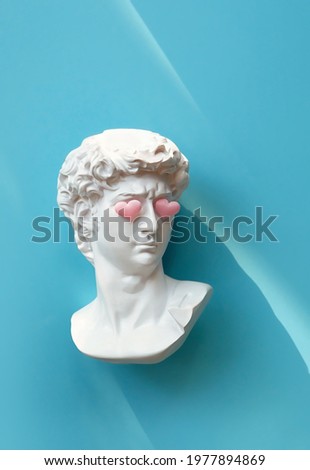 Plaster head of David with pink hearts on eyes on blue background. White sculpture of antique head, modern style. creative minimal art concept. symbol of love, romantic holiday, Valentine's Day