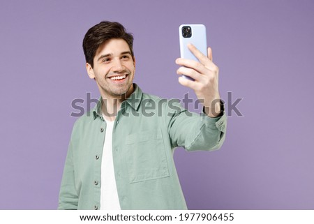 Young fun smiling caucasian man 20s in mint shirt white t-shirt doing selfie shot on mobile phone post photo on social network isolated on purple background studio portrait. People lifestyle concept.
