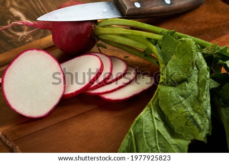 Fresh red sliced radish on a wooden board with the knife on side. On whole radish with the green leaves.                               