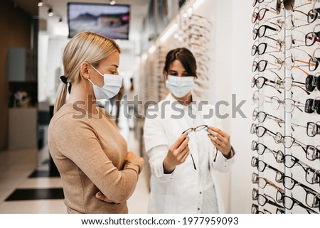 Beautiful woman choosing eyeglasses frame in modern optical store. Female seller specialist helps her to make right decision. They are wearing protective face mask against virus pandemic. Royalty-Free Stock Photo #1977959093