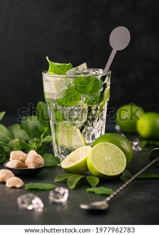 Glass of Mojito cocktail with ice cubes and disk stirrer,mint and lime on black board with spoon and fresh limes with cane sugar. Best party drink. Royalty-Free Stock Photo #1977962783