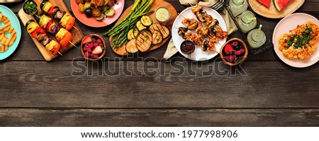 Healthy plant based summer bbq top border. Above view over a dark wood banner background. Grilled fruit and vegetables, skewers, cauliflower steak and vegetarian sides. Copy space.